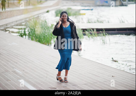 African American dark scuoiati plus modello dimensioni poste in un azzurro vestito lucido e nero pelliccia contro il lago e parlando dal telefono cellulare. Foto Stock