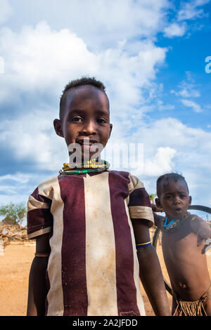 Turmi, Etiopia - Nov 2018: Hamer tribù kid in posa striped t-shirt e capretto con pneumatico giocattolo in retro. Valle dell'Omo Foto Stock