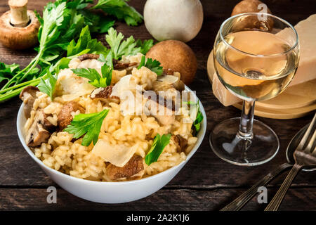 Risotto ai funghi, funghi Riso, con ingredienti e un bicchiere di vino bianco al buio su un rustico sfondo di legno Foto Stock