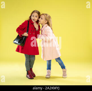 Una piena lunghezza Ritratto di un luminoso alla moda ragazze caucasiche in un impermeabile tenendo un sacco nero su giallo di sfondo per studio. Autunno e primavera moda per bambini. Carino elegante ragazza bionda. Foto Stock