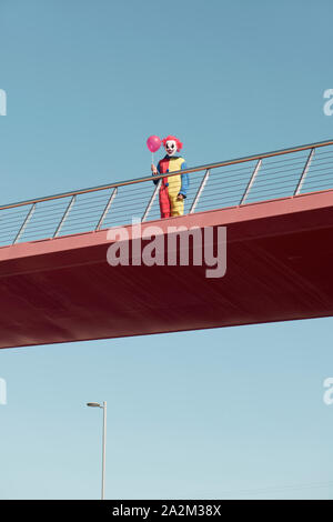 Un inquietante clown indossa un colorato di giallo, rosso e blu costume, tenendo in mano un palloncino rosso in mano in piedi in un ponte all'aperto Foto Stock