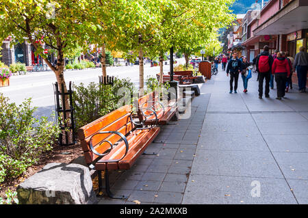 Panche di legno e alberi che fiancheggiano il marciapiede con gente che cammina lungo i negozi su Banff Avenue in caduta, Banff, Alberta, Canada Foto Stock