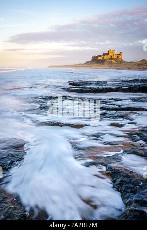 Il castello di Bamburgh è illuminato dal basso sole serale come rulli enorme crash sulla spiaggia al di sotto di produrre schiuma percorsi e modelli ad alta marea. Foto Stock