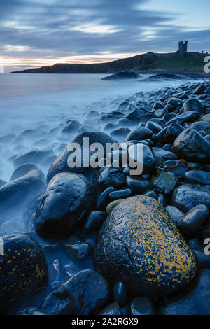 Le imponenti rovine del castello di Dunstanburgh torre sopra il dolerite ciottolosa base Embleton Bay come alba luce inizia a evidenziare la scena. Foto Stock