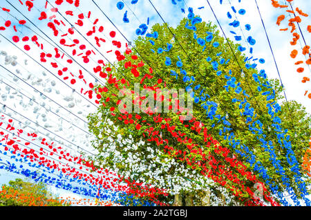 Bella strada colorato decor in Loreto, Madeira, Portogallo. Celebrazione della festa religiosa. Rosso, bianco, blu e arancio fiori di carta appeso in aria. Albero verde in background. Foto Stock