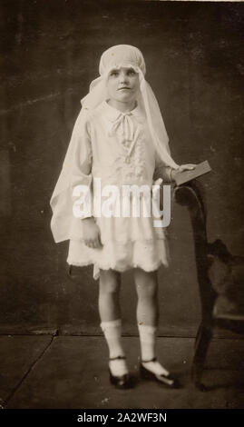 Fotografia digitale - Cartolina, ragazza vestita per la prima volta la Santa Comunione, Williamstown, 1929, Studio ritratto della ragazza vestita per la prima comunione, in un abito bianco e il velo. Il suo velo è mantenuta attorno alla sua testa come un cappuccio con nastro. Ella è in piedi da una sedia a destra e portante un libro in mano. Questa fotografia mostra Margaret Hogarty, vestito per la sua Prima Comunione, in Williamstown, 1929. La sua Prima Comunione ha avuto luogo a St Mary's chiesa cattolica Williamstown. Ella era Foto Stock