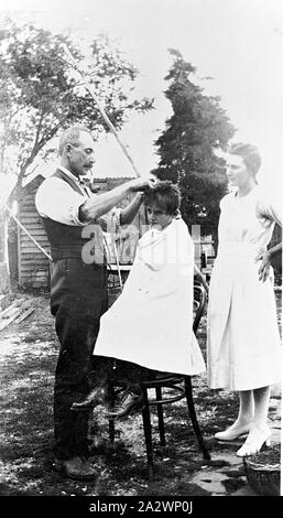 Negativo - Ballarat, Victoria, pre 1930, un uomo sta dando un giovane ragazzo in un taglio di capelli. Il ragazzo è seduto su una sedia in un cortile. Una donna Orologi Foto Stock