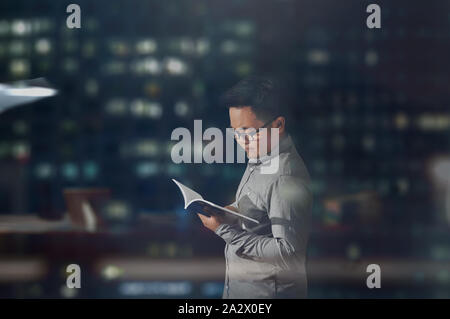 Una vista laterale colpo di un giovane uomo a maniche lunghe e bicchieri pacificamente la lettura di un libro all'interno di un ufficio scuro stanza a notte tempo. Foto Stock