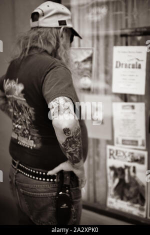 Vista posteriore dei lunghi capelli straggly uomo di mezza età che indossa cappello da baseball e il coltello sulla cinghia e il braccio tatuaggi storefront studi contemplati nel poster Foto Stock