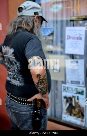 Vista posteriore dei lunghi capelli straggly uomo di mezza età che indossa cappello da baseball e il coltello sulla cinghia e il braccio tatuaggi storefront studi contemplati nel poster Foto Stock