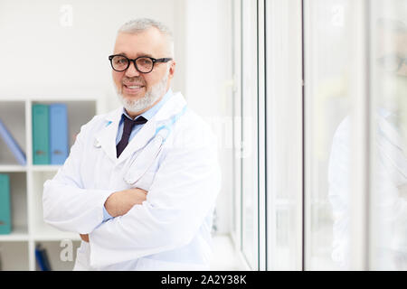 Ritratto di senior medical professional in occhiali e in camice bianco in piedi con le braccia incrociate e sorridente alla fotocamera in ospedale Foto Stock