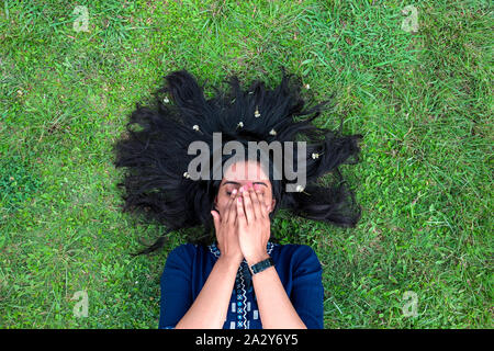 Giovane e bella ragazza distesa sul prato con capelli sparsi che copre il volto con le mani Foto Stock