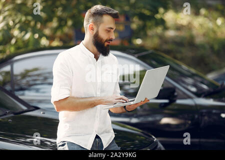 Uomo bello in una camicia bianca. Imprenditore lavora con il computer portatile. Uomo in piedi vicino a un auto Foto Stock
