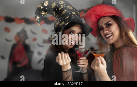 Due donne asiatiche per il make-up e vestiti in costumi di Halloween, in piedi tenendo un bicchiere di rosso e arancio acqua con poca velatura, che puntano il dito di un Foto Stock