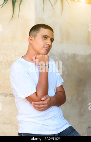 Ritratto di Allegro adolescente all'aperto contro un muro bianco sfondo, closeup. Ritratto di pensieroso piacevole guardando giovane adolescente di sesso maschile, mantiene la mano sotto il mento e guarda pensieroso in avanti . Foto Stock