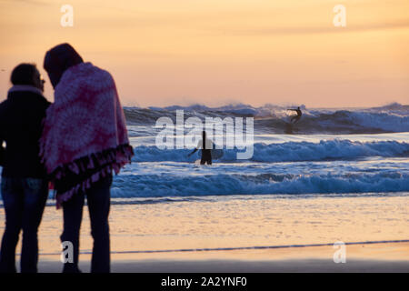 Un giovane sulla spiaggia sabbiosa e surfisti cavalcare le onde durante il tramonto a Santa Cruz, in California. Foto Stock