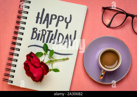 Happy Birthday greeting card - scrittura in un taccuino, laici piatta con caffè, red rose e occhiali di protezione Foto Stock