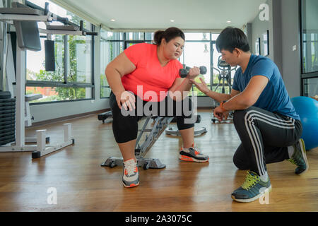 Due Asian trainer uomo e donna sovrappeso che esercitano con il manubrio insieme in una palestra moderna, felice e sorridere durante gli allenamenti. Grasso donne prendersi cura di hea Foto Stock