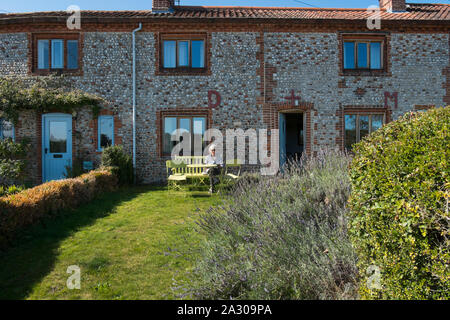 Capelli grigi nonna seduta su una panchina leggendo una rivista al di fuori della Hall Wiveton holiday cottages in Marsh Lane, Wiveton, North Norfolk, Regno Unito Foto Stock