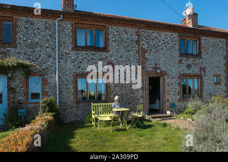 Capelli grigi nonna seduta su una panchina leggendo una rivista al di fuori della Hall Wiveton holiday cottages in Marsh Lane, Wiveton, North Norfolk, Regno Unito Foto Stock