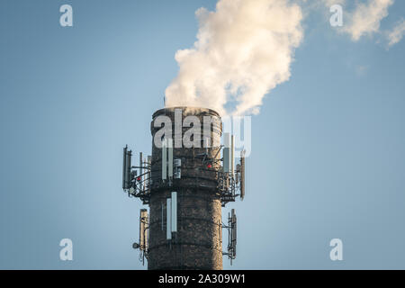 In alto di alto camino industriale con più telefono cellulare antenne, fumo comming out sul cielo azzurro sfondo Foto Stock