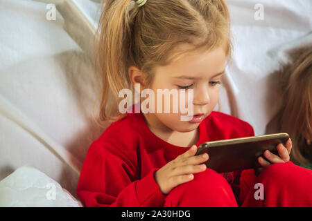 Bambina in morbido e caldo pigiama a giocare a casa. Bambini caucasici in abiti colorati avendo divertimento. Infanzia, comfort di casa, la felicità. Seduti sul divano e giocare con lo smartphone o l'apprendimento. Foto Stock