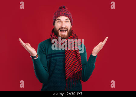 Wow, non so gesto di un uomo barbuto che indossa inverno, sciarpa e cappello su sfondo rosso. Foto Stock