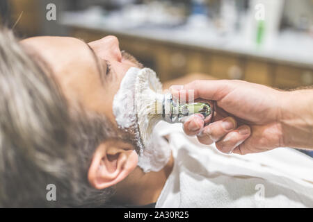 Parrucchiere professionale applicazione di schiuma da barba sulla pelle del client in barberia. Foto Stock