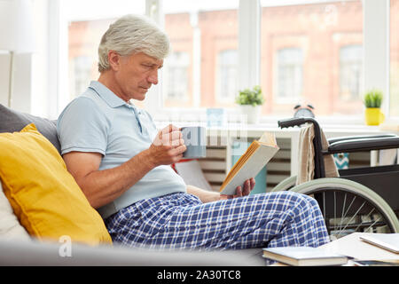 Senior uomo in vestiti domestici in appoggio sul divano con la tazza di caffè e la lettura di un libro nel salotto di casa Foto Stock