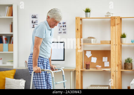 Uomo maturo in piedi e cercando di camminare con l'aiuto di walker in camera a casa Foto Stock