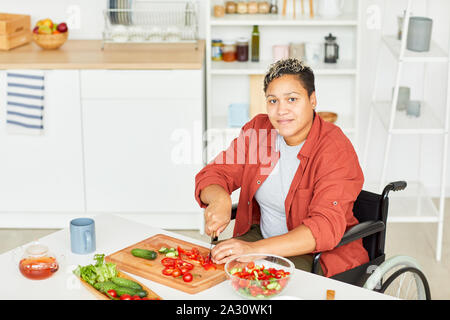 Ritratto di disabili africani giovane donna con capelli corti guardando la fotocamera mentre è seduto nella sedia a rotelle e tagliare le verdure in cucina Foto Stock