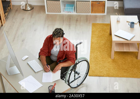 Giovane donna africana seduto nella sedia a rotelle al suo posto di lavoro e di rendere note in un documento di Office Foto Stock