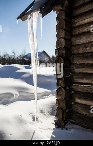 Un potente, trasparente lungo ghiacciolo pende dal bordo del tetto della vecchia casa di log e raggiunge il cumulo di neve. In controluce del sole, Foto Stock