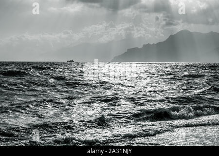 Bianco e nero seascape con singola nave e montagne silhouette sul bellissimo cielo nuvole sfondo con raggi solari Foto Stock