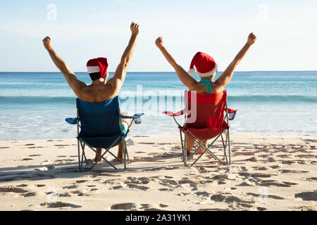 Vista posteriore di una giovane coppia con cappelli di Babbo Natale seduto sulla sedia alzando le braccia Foto Stock