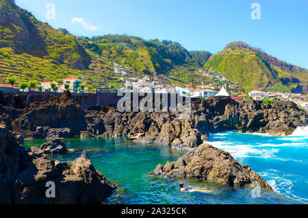 Donna di nuoto in piscine naturali nell'Oceano Atlantico, l'isola di Madeira, Portogallo. Composta di roccia vulcanica, nel mare che fluisce naturalmente. Villaggio Porto Moniz sulla collina rocciosa in background. Foto Stock