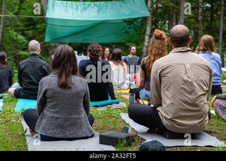 Un gruppo multigenerazionale di persone sono visti da dietro, seduti su Materassini da yoga durante un rituale sciamaniche nel bosco, in cerca di illuminazione e consapevolezza. Foto Stock