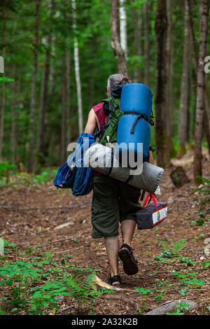 Un uomo di mezza età è visto a piedi attraverso i fitti boschi che trasportano zaini, una tenda e attrezzature per il campeggio, in cerca di una paura di massa per memore di ritiro. Foto Stock