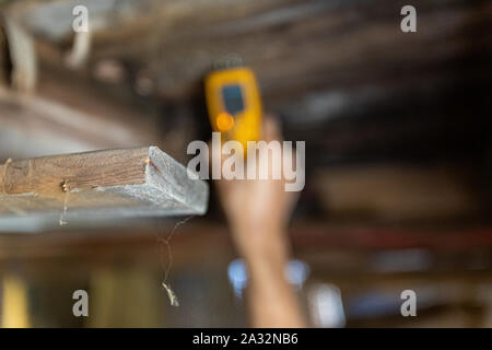 Una vista ravvicinata su un vecchio e polveroso listone di legno all'interno di una casa, una mano sfocata è visto in possesso di un rilevatore di umidità durante un ambiente chiuso l'esame. Foto Stock