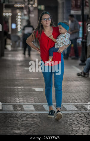 Una singola donna che indossa una maglietta rossa e blu jeans sta portando la sua figlia mentre si cammina su una strada pulita. Foto Stock