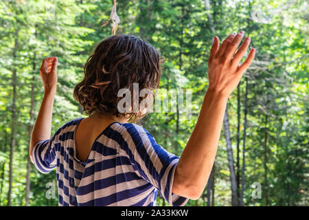 Una vista ravvicinata di una giovane donna con spalla Ricci Lunghezza capelli e a strisce blu top, in piedi con le mani alzate in aria durante un consapevole esercizio di meditazione. Foto Stock