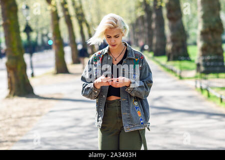 Giovane donna urbano con moderni acconciatura utilizza lo smartphone a piedi in strada in un parco urbano. Foto Stock