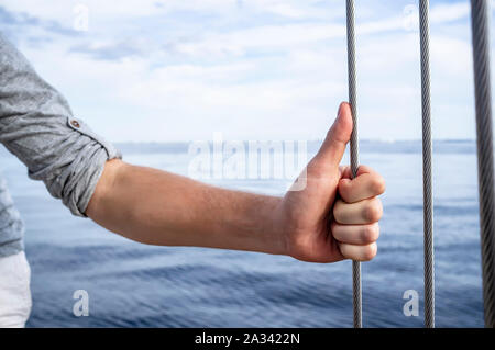 L'uomo tiene per mano con 'pollice in alto' sul metallo guy sullo yacht, contro lo sfondo del mare, le onde e il cielo di nuvole, su una calda estate Foto Stock