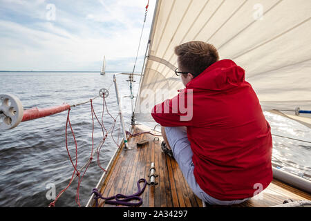 Un Uomo in camicia rossa e i bicchieri si siede sul ponte di una barca in movimento e appare in lontananza, contro lo sfondo del mare, al cielo nel cl Foto Stock