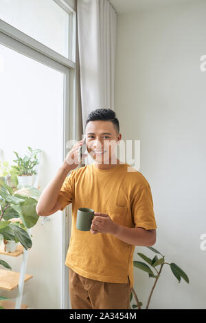Giovane uomo che indossa abiti casual a parlare su un telefono cellulare al mattino in corrispondenza di una finestra Foto Stock