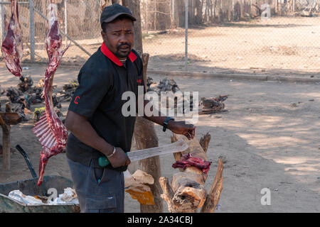 Opuwo, Namibia - 25 Luglio 2019: uomo nero macellazione carne di capra a un mercato tradizionale con carne appesi in background in Opuwo, Namibia Foto Stock