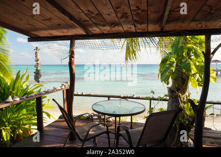 Vista di un azzurro laguna tropicale dal ponte di legno di un bungalow con un tavolo e sedie e incorniciato da palme e conchiglie Foto Stock