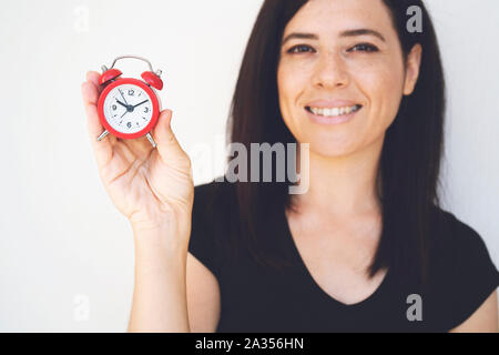 Felice giovane donna in nero t-shirt una holding sveglia rossa e agitando la mano isolato su bianco Foto Stock