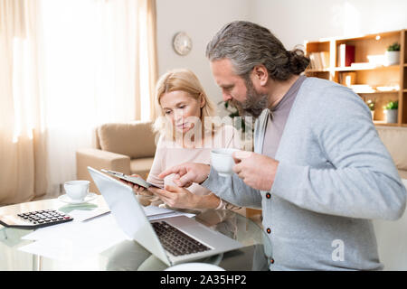 Maturo uomo barbuto con tazza di tè puntando al documento detenuto dalla moglie Foto Stock