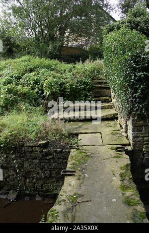 Vecchia lastra di pietra con raccordi in metallo la formazione di ponte su streem con gradini in pietra e il percorso da siepe e arbusti in Yorkshire Inghilterra Foto Stock